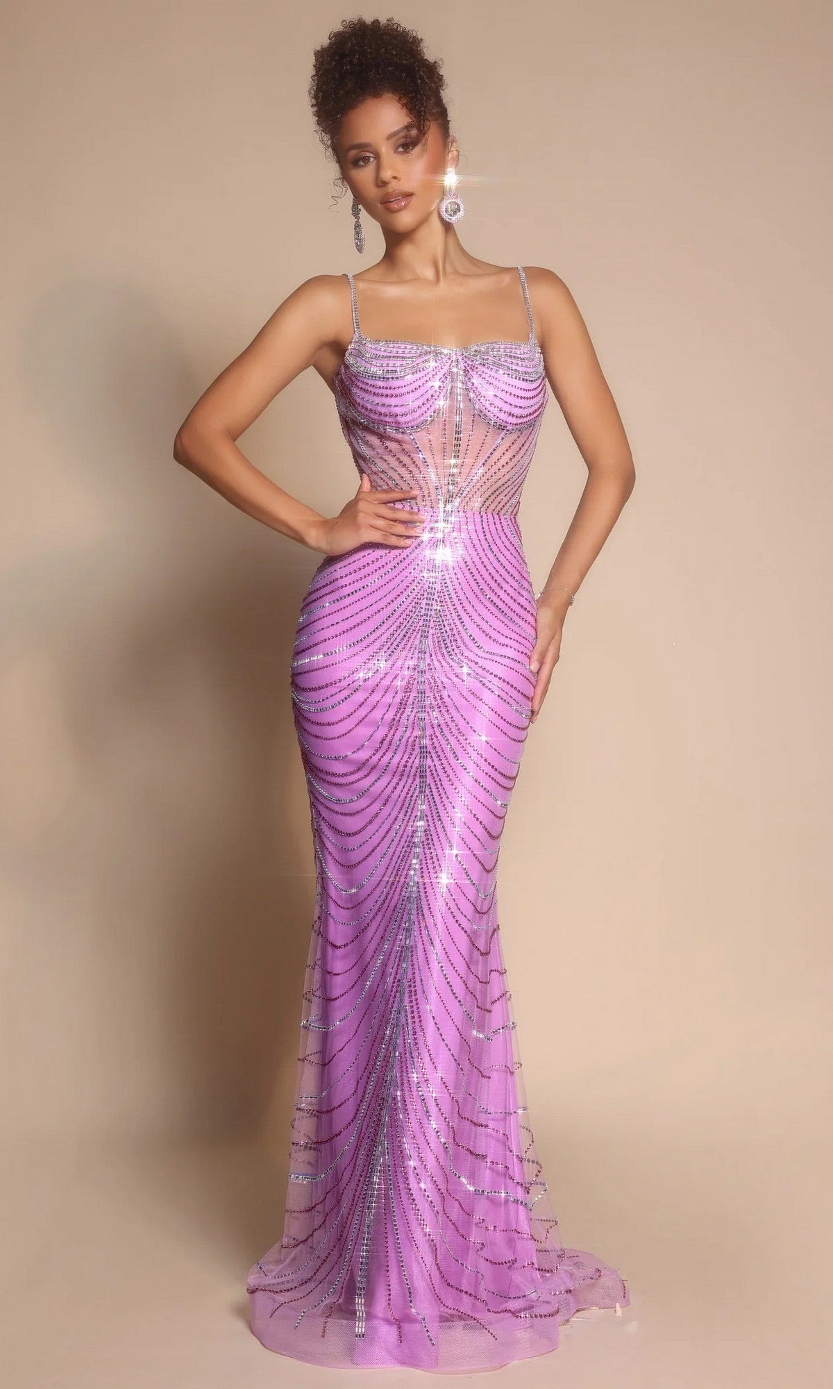A woman poses in the Portia and Scarlett PS26095 long formal dress, fitted and floor-length with thin straps and rhinestone embellishments, shimmering in lilac against a plain beige background.